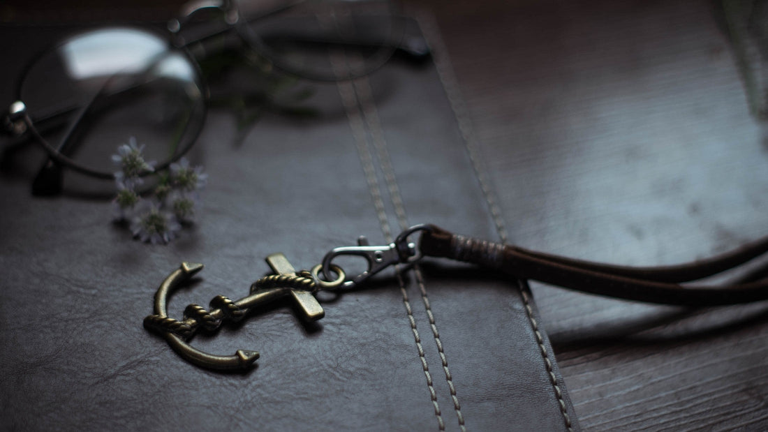 Close-up of a bronze anchor keychain on dark brown, double-stitched leather, with blurred glasses and small flowers in the background—highlighting designer craftsmanship.