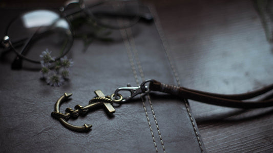Close-up of a bronze anchor keychain on dark brown, double-stitched leather, with blurred glasses and small flowers in the background—highlighting designer craftsmanship.