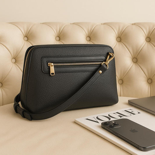 Black leather handbag with gold zipper on a beige surface with a magazine and phone.