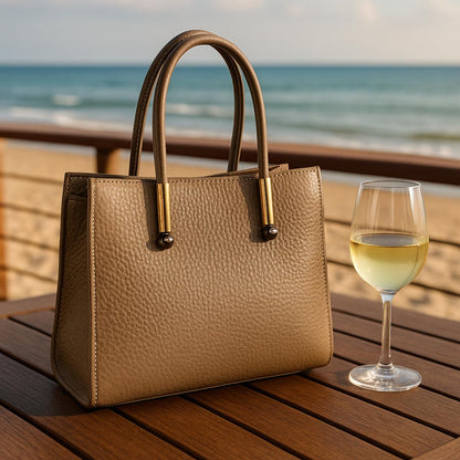 Beige handbag on a wooden table with a glass of white wine by the ocean.